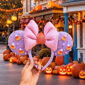 May include: A pair of lavender-colored headband ears featuring a large, textured pink bow in the center. The ears are decorated with small, scattered candy corn motifs and tiny pearl-like beads. The headband is covered in a matching lavender fabric.