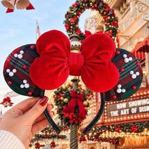 May include: A headband featuring two circular ears with a dark green and red plaid pattern, accented by white pearl Mickey-shaped icons and small red beads. A large, plush, bright red velvet bow is centered between the two ears.