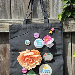 May include: A black canvas tote bag decorated with various circular pin-back buttons and artificial flowers. The buttons feature text including faith, grace, Jesus is king, and Love keeps no record of wrongs. Large peach and pink fabric flowers are attached.