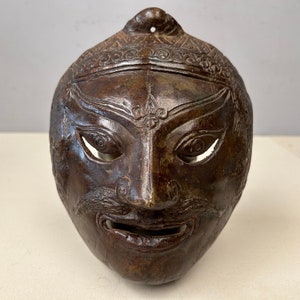 Puede incluir: Una máscara de bronce marrón oscuro con detalles intrincados, que incluye una cara tallada con una boca ancha y ojos, y una parte superior decorativa. La máscara es probablemente de una cultura asiática tradicional.