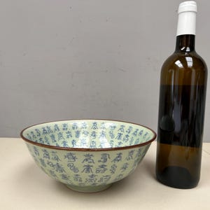 May include: A light green ceramic bowl with blue Chinese characters and a brown rim sits next to a brown wine bottle with a white label. The bowl has a wide, shallow shape.