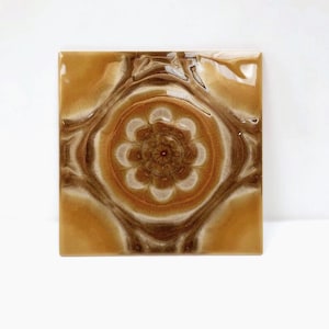 Puede incluir: Un azulejo de cerámica cuadrado con un patrón de remolino marrón y blanco. El centro del azulejo presenta un diseño de flor blanco con un centro marrón.
