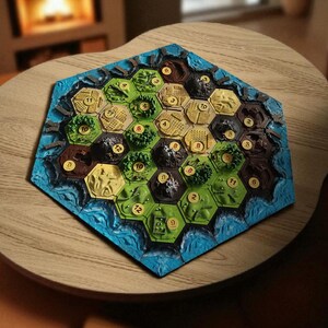 May include: A hexagonal board game with terrain tiles, including green fields, brown mountains, and blue water. The game board is set on a wooden table, with a fireplace in the background. The tiles have numbers.