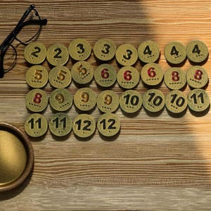 May include: A collection of small, round, antique-style game tokens with numbers from 2 to 12. The tokens are arranged on a wooden surface, with a pair of eyeglasses and a small brass tray visible.