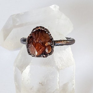 May include: A handcrafted ring featuring a raw, amber-colored gemstone set in a dark, oxidized metal band. The gemstone has a unique, irregular shape, and the ring is displayed on a clear crystal, showcasing its natural beauty.