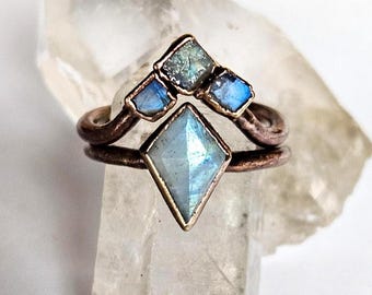 May include: A copper-toned ring with a geometric design. The ring features a large, light blue, diamond-shaped stone at the center, with three smaller, blue-green stones above. The ring is set against a clear, crystalline background.