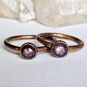 May include: Two copper-toned rings with faceted, round, purple gemstones. The rings are set on a white surface, with a blurred background. The gemstones have a subtle sparkle, and the metal has a rustic finish.