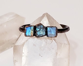 May include: A handcrafted ring with a dark metal band, featuring three square-cut, iridescent blue gemstones. The gemstones have a rough, natural texture. The ring is displayed on a clear crystal, with another crystal in the background.