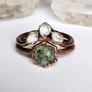 May include: Two copper rings. The top ring features three clear, irregular-shaped stones. The bottom ring has a hexagonal green stone with a mossy appearance. The rings are set against a white background.
