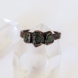 May include: A copper-toned ring featuring three raw, green gemstones. The ring's band has a textured appearance, and the stones are set in a way that highlights their natural, irregular shapes. The ring is displayed on a clear, crystal-like surface.
