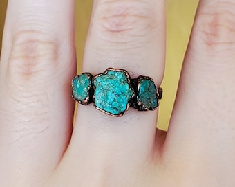 May include: A copper-toned ring featuring three raw, turquoise-colored stones. The stones are irregularly shaped and set in a simple, rustic band. The ring is displayed on a hand, showcasing the natural color and texture of the gemstones.