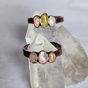 May include: Two copper-toned rings, each featuring three oval opals. The opals display a range of colors, including pink, green, and white. The rings are displayed on a clear, crystalline surface, showcasing the jewelry's design and color variations.