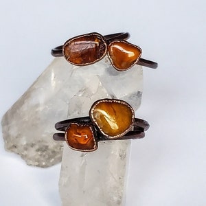 Op de afbeelding: Twee handgemaakte ringen met bruine metalen banden en onregelmatig gevormde, amberkleurige stenen. De stenen hebben een gepolijste, glanzende uitstraling. De ringen worden gepresenteerd op een witte achtergrond met een kristalachtig object.