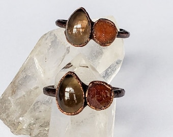 May include: Two copper rings, each featuring a teardrop-shaped, translucent, brown gemstone paired with a smaller, round, reddish-brown stone. The rings are displayed on a clear, crystalline surface against a white background.