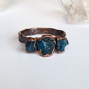 May include: A handcrafted ring featuring three raw, teal-colored gemstones set in a textured, dark copper band. The gemstones are irregularly shaped, adding to the ring's unique, natural aesthetic. The ring is displayed on a white surface.