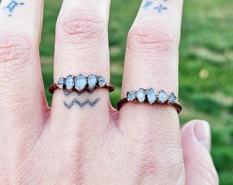 May include: Two copper-colored rings, each featuring a row of light blue, irregular-shaped stones. The rings are displayed on fingers with tattoos, set against a blurred green background.
