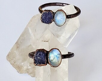 May include: Two handcrafted rings with dark metal bands. Each ring features a rough-cut, dark blue gemstone paired with a smooth, oval-shaped, light blue gemstone. The rings are displayed on a clear, crystalline surface.