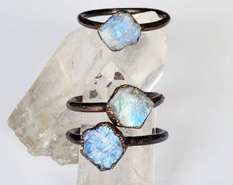 May include: Three rings with raw, iridescent moonstone gemstones set on dark metal bands. The moonstones display shades of blue, white, and silver. The rings are displayed on a clear crystal.
