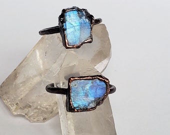 May include: Two rings with a raw, rectangular, blue-hued stone set in a dark metal band. The stones have a natural, irregular shape with a shimmering, iridescent quality. The rings are displayed on a clear, crystalline surface.