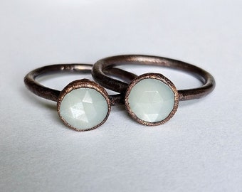 May include: Two rings with a rustic, dark brown band and a light blue, faceted gemstone. The gemstones are set in a copper-colored bezel. The rings are displayed on a white surface.