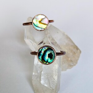 May include: Two rings featuring circular abalone shell inlays set in copper bands. The top ring displays iridescent white, pink, and green tones, while the bottom ring showcases deep blue, green, and black hues with a polished, reflective finish.