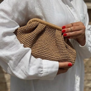 May include: A brown woven clutch bag with a gathered design. The bag is held in a hand with red nail polish.