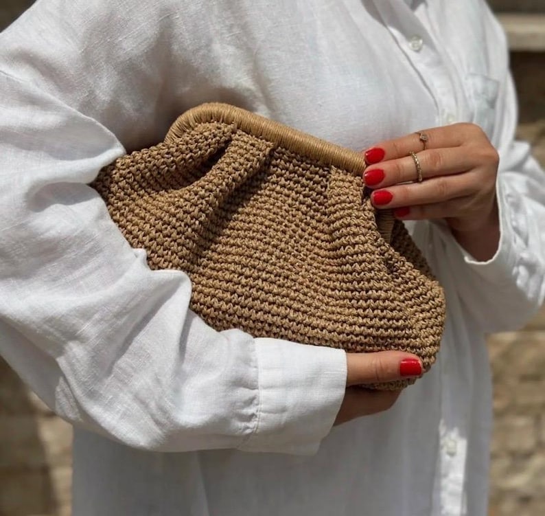 May include: A brown woven clutch bag with a gathered design. The bag is held in a hand with red nail polish.