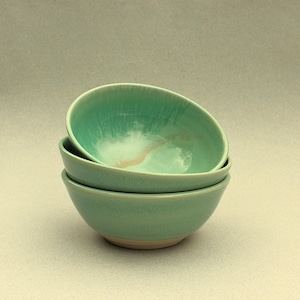 May include: Two teal ceramic bowls stacked on top of each other. The bowls have a glossy finish and a simple, rounded shape. The interior of the top bowl has a lighter, almost white, area with a hint of tan. The bowls have a natural, unglazed base. The bowls are handmade.