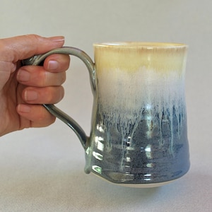 May include: A ceramic mug featuring a two-tone glaze with a creamy beige top that transitions into a deep navy blue base. The glaze exhibits a vertical drip pattern where the colours blend, and the mug includes a curved handle on the side.