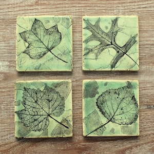 May include: Set of four square ceramic coasters with leaf imprints. Each coaster features a different leaf design in black against a mottled green background. The coasters are approximately 10 cm square.