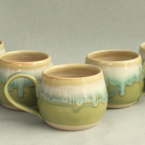 May include: A set of four ceramic mugs with a rounded shape and a handle on the side. Each mug features a two-tone glaze with a light beige or cream colour on the upper portion and a sage green colour on the lower portion, with a drip effect.