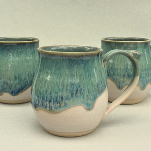 May include: Three ceramic mugs with a teal glaze over a cream base. The mugs have rounded bodies, large handles, and a dripped glaze effect. The mugs are handmade and suitable for coffee or tea.