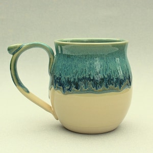 May include: A handcrafted ceramic mug with a rounded form. The upper section is glazed in teal and blue hues, featuring a dripped design. The lower part is a creamy beige. The handle mirrors the teal and beige tones. Ideal for hot drinks, this mug is unique.