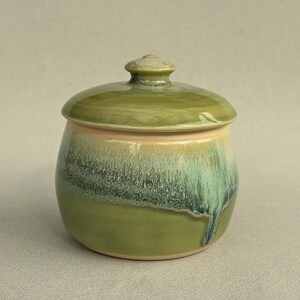 May include: A ceramic jar with a lid, featuring a green glaze. The jar has a rounded body with a lid and a small knob. The glaze transitions from light to dark green, creating a unique, handcrafted appearance.