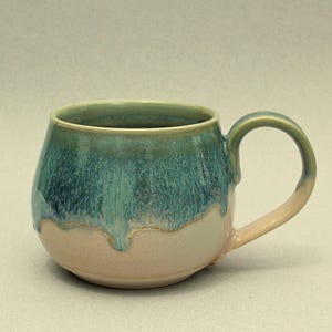 May include: A handcrafted ceramic mug with a rounded body and a large handle. The mug has a colour gradient, with a teal and green glaze on the upper half that drips down to a light pink base. The interior is a light green.