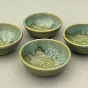 May include: Four handmade ceramic bowls. Each bowl is a muted green with a turquoise and white glaze around the rim. The interior of each bowl has a spiral design. The bowls are arranged in a group.