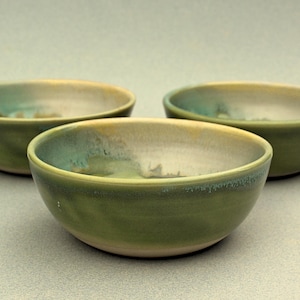 May include: Three handmade ceramic bowls with a green and turquoise glaze. The bowls have a rounded shape and a smooth, glossy finish. The interior of the bowls is a light beige colour.