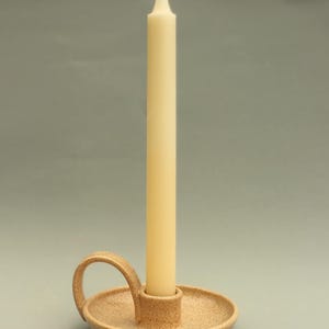May include: A beige speckled ceramic candle holder with a circular base and a curved side handle. A single, tall, off-white taper candle is inserted into the central cylindrical socket of the holder, standing upright against a neutral background.
