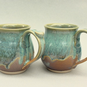 May include: Two handcrafted ceramic mugs with a teal and brown glaze. The mugs have rounded bodies, sturdy handles, and a light-coloured rim. The glaze creates a marbled effect, adding visual interest. These mugs are ideal for enjoying coffee, tea, or other beverages.