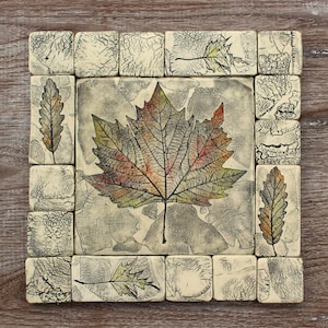 May include: A ceramic tile wall art piece featuring a large, detailed maple leaf in shades of brown, yellow, and red. The leaf is surrounded by smaller leaves in a similar colour scheme, all set against a cream-coloured background.