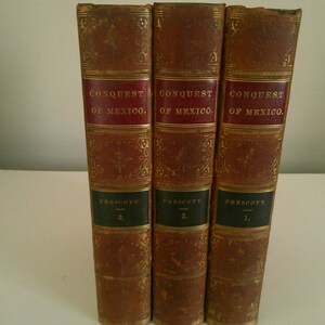 Puede incluir: Tres libros antiguos titulados "Conquista de México" de Prescott. Los libros tienen cubiertas de cuero marrón con detalles dorados y etiquetas de título rojas y negras. Los libros están numerados 1, 2 y 3.