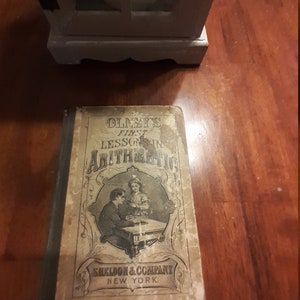 Puede incluir: Un libro antiguo titulado "Olney's First Lessons in Arithmetic" con una ilustración en blanco y negro de dos niños sentados en un escritorio. El libro está publicado por Sheldon & Company en Nueva York.