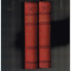 Puede incluir: Dos libros rojos antiguos titulados "Memorias de Napoleón" de la Duquesa D'Abrantès. Cada libro tiene letras doradas y un diseño de estrella con la letra "N" en el lomo. Los volúmenes I y II son visibles.