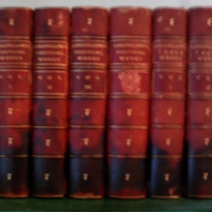 Puede incluir: Un conjunto de siete libros encuadernados en cuero rojo con letras doradas. Los libros se titulan "Longfellow's Poetical Works" y cada libro tiene un número de volumen.