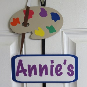 May include: A brown paint palette with colorful paint splatters, a paintbrush, and a sign that says "Annie's 5th" hanging on a white door.
