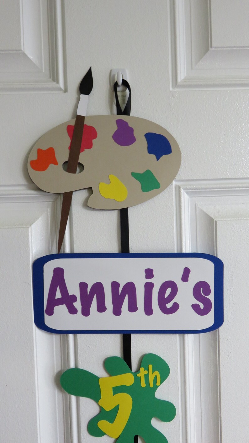 May include: A brown paint palette with colorful paint splatters, a paintbrush, and a sign that says "Annie's 5th" hanging on a white door.