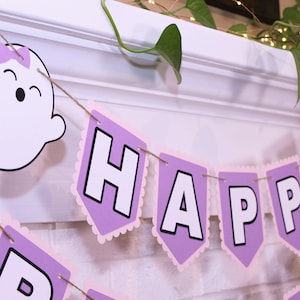 May include: A festive banner with the words "HAPPY BIRTHDAY" in white letters on purple pennants. The banner is strung with twine and features a smiling ghost with a purple bow. The background is a white brick wall.
