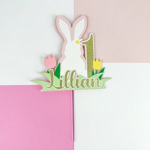 May include: A white bunny cake topper with a pink and green grass base. The bunny has a yellow flower on its head. The number 1 is gold glitter and the name Lillian is written in gold glitter on the base.