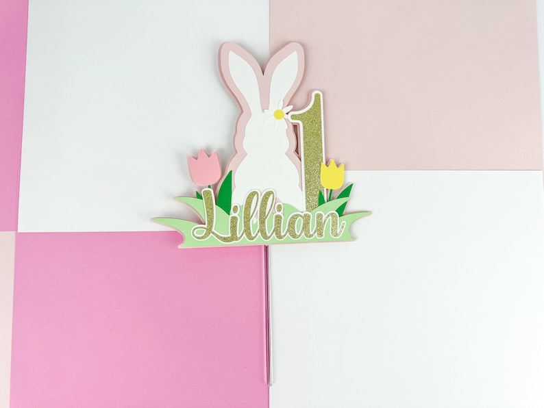 May include: A white bunny cake topper with a pink and green grass base. The bunny has a yellow flower on its head. The number 1 is gold glitter and the name Lillian is written in gold glitter on the base.