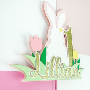 May include: A white bunny cake topper with a pink flower, green leaves, and a gold glitter number one. The name "Lillian" is written in gold glitter on a green background.